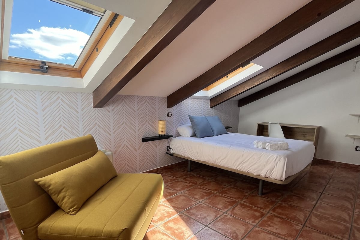 A bedroom with a bed, a sofa, a nightstand, and a skylight.