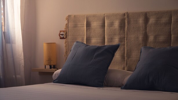 Una cama bien hecha con dos almohadas, una lámpara de noche y un reloj de pared.