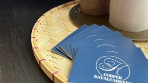 Un logotipo con 'CORPER NAVALCARNERO' y dos velas encendidas sobre una bandeja tejida.