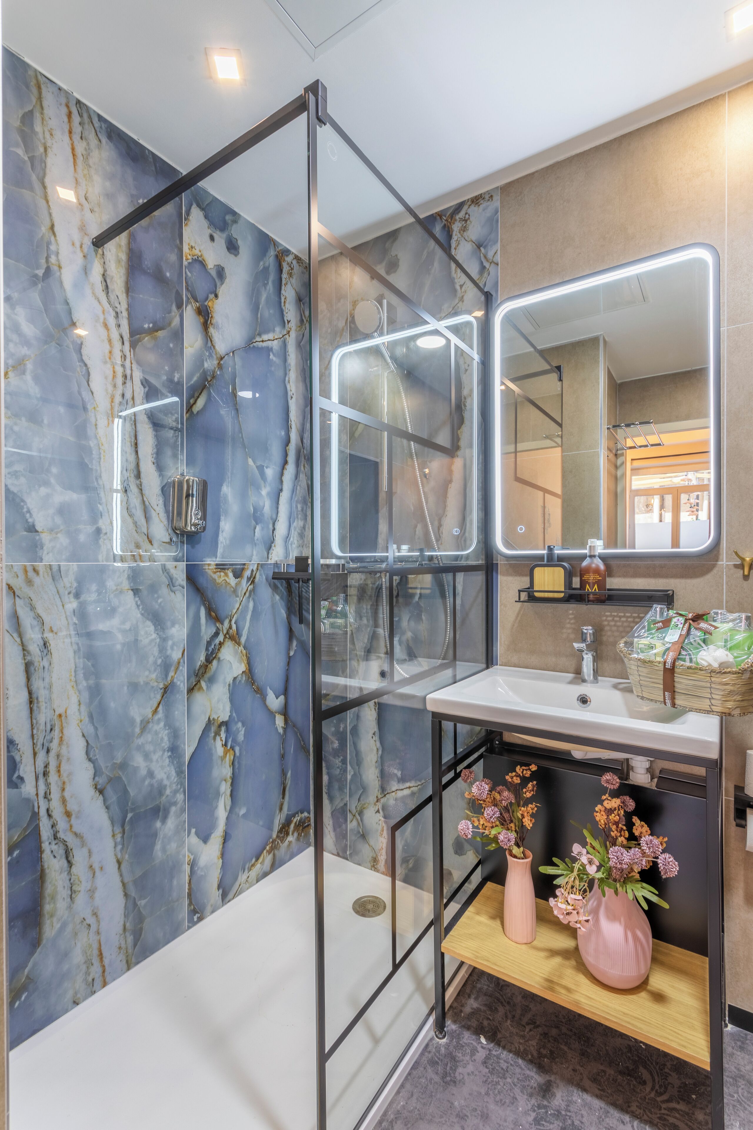 A modern bathroom with a glass shower enclosure, marble walls, a white sink, and two pink vases on a wooden shelf.