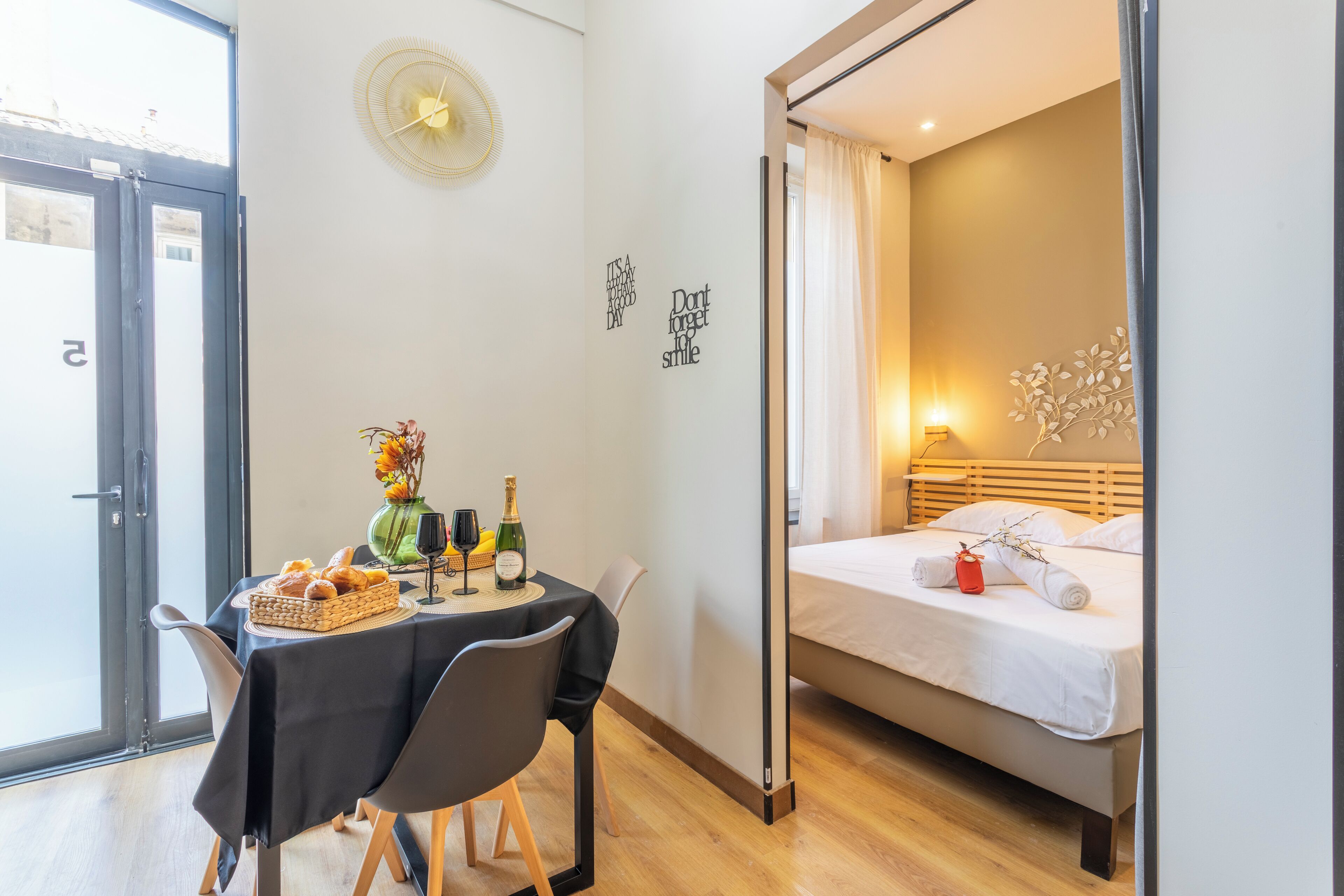 A hotel room with a bed, a table with breakfast, a vase with flowers, and motivational wall art.