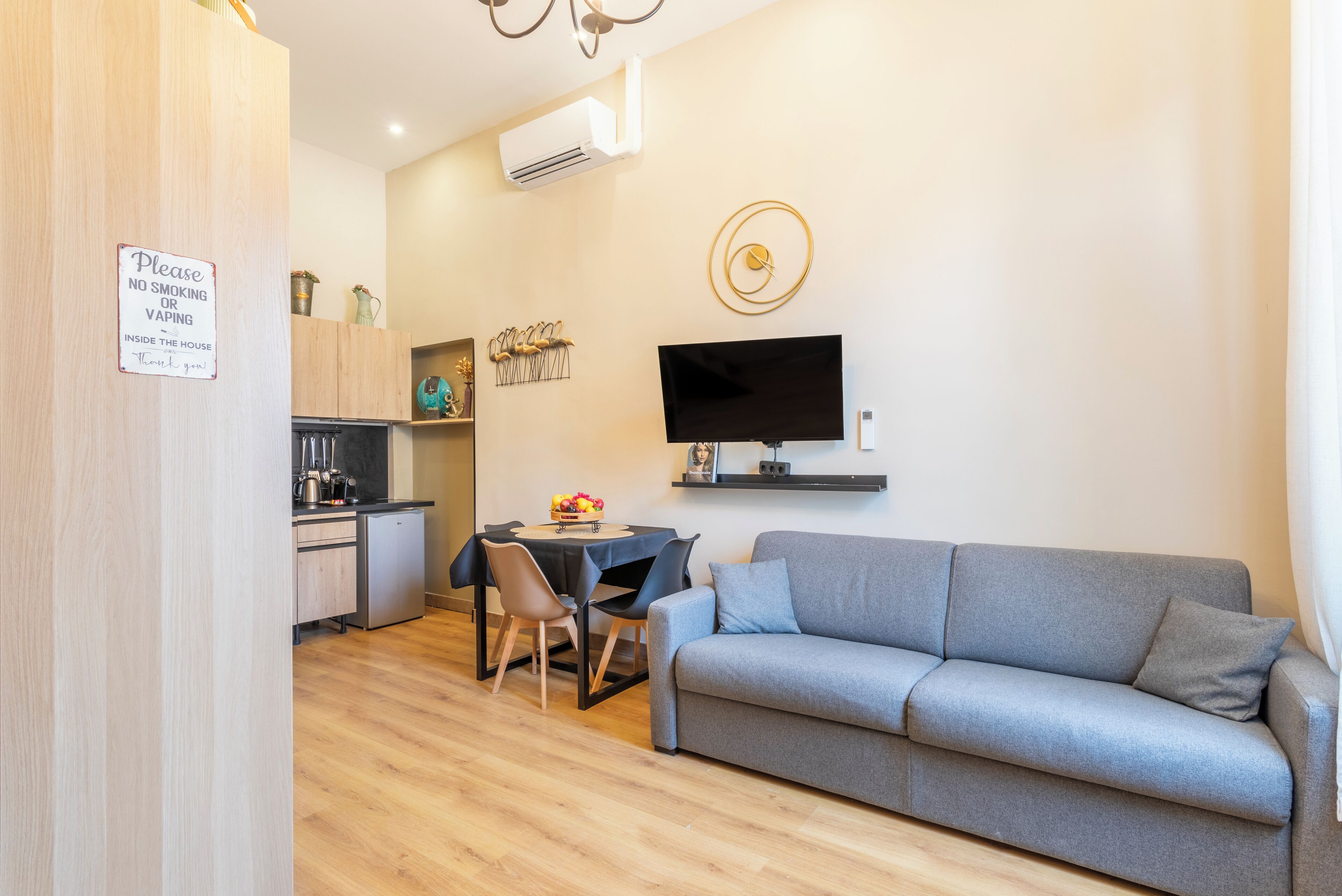 Een moderne woonkamer met een grijze bank, een eettafel met stoelen, een televisie en een keukenhoek met een bordje waarop roken en dampen verboden is.