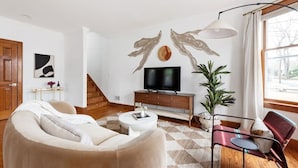 A modern living room with a beige sofa, a white coffee table, a TV on a wooden stand, and a large window with curtains. - Espresso Martini (Atlanta)