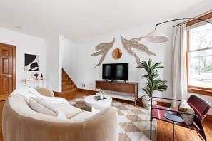 Una sala de estar moderna con un sofá color beige, una mesa de centro blanca, un televisor sobre un mueble de madera y un amplio ventanal con cortinas.