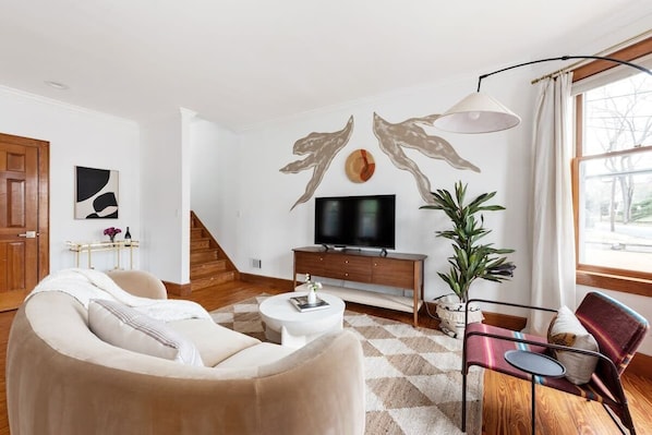 A modern living room with a beige sofa, a white coffee table, a TV on a wooden stand, and a large window with curtains.