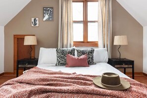 A bedroom with a bed, two nightstands, a window with curtains, and a wall with two framed pictures.