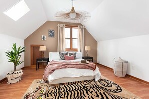 Une chambre avec un plafond en pente, un lit surmonté d’un tapis à motifs zébrés, une plante en pot et un luminaire suspendu.