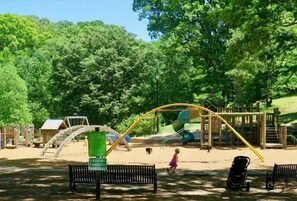 Une aire de jeux avec des balançoires, un toboggan et un espace sablonneux, le tout entouré d’arbres.