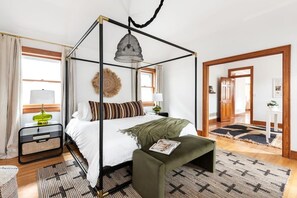Un lit à baldaquin avec un luminaire suspendu, une table de chevet avec une lampe, un banc vert et un tapis à motifs.