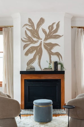 A living room with a fireplace, a large wall art piece, a round ottoman, and two armchairs.