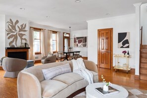 A living room with a beige sofa, a coffee table with books and flowers, a fireplace, and a dining area with a table and chairs.