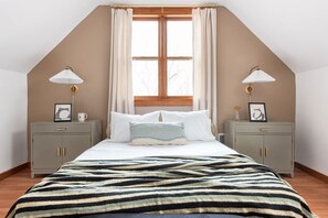 Une chambre avec un plafond en pente, une grande fenêtre avec des rideaux, un lit recouvert d’une couverture à rayures, deux tables de chevet avec des lampes et des tableaux encadrés, et une armoire grise.