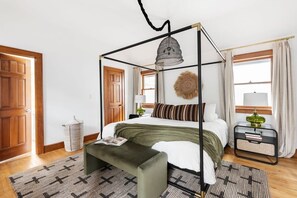 A four-poster bed with a green upholstered bench, a patterned rug, and a bedside table with a lamp.