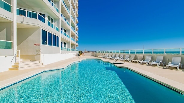 Un espace piscine avec une eau d’un bleu limpide, un bâtiment blanc doté de balcons et une vue sur l’océan.