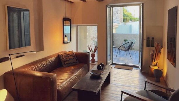 A cozy living room with a leather sofa, a coffee table, and a dining table. There is a large window with a view of the outside. - Spacieux 2 Chambres Dans le Centre D'arles Avec Terrasse (Arles)