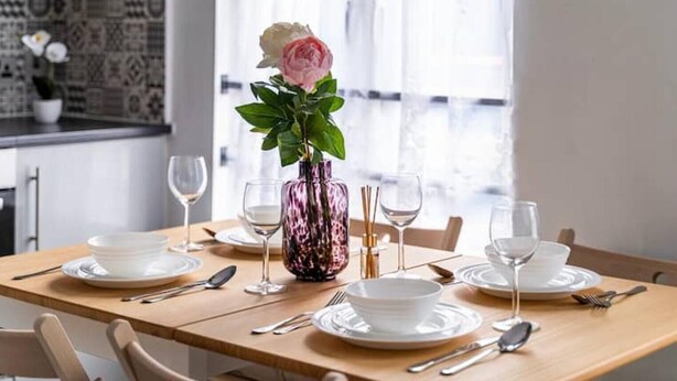 Et spisebord dækket med hvide tallerkener, bestik og en pink vase med en hvid rose.