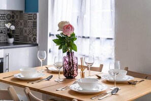 A dining table set with white plates, silverware, and a pink vase with a white rose.