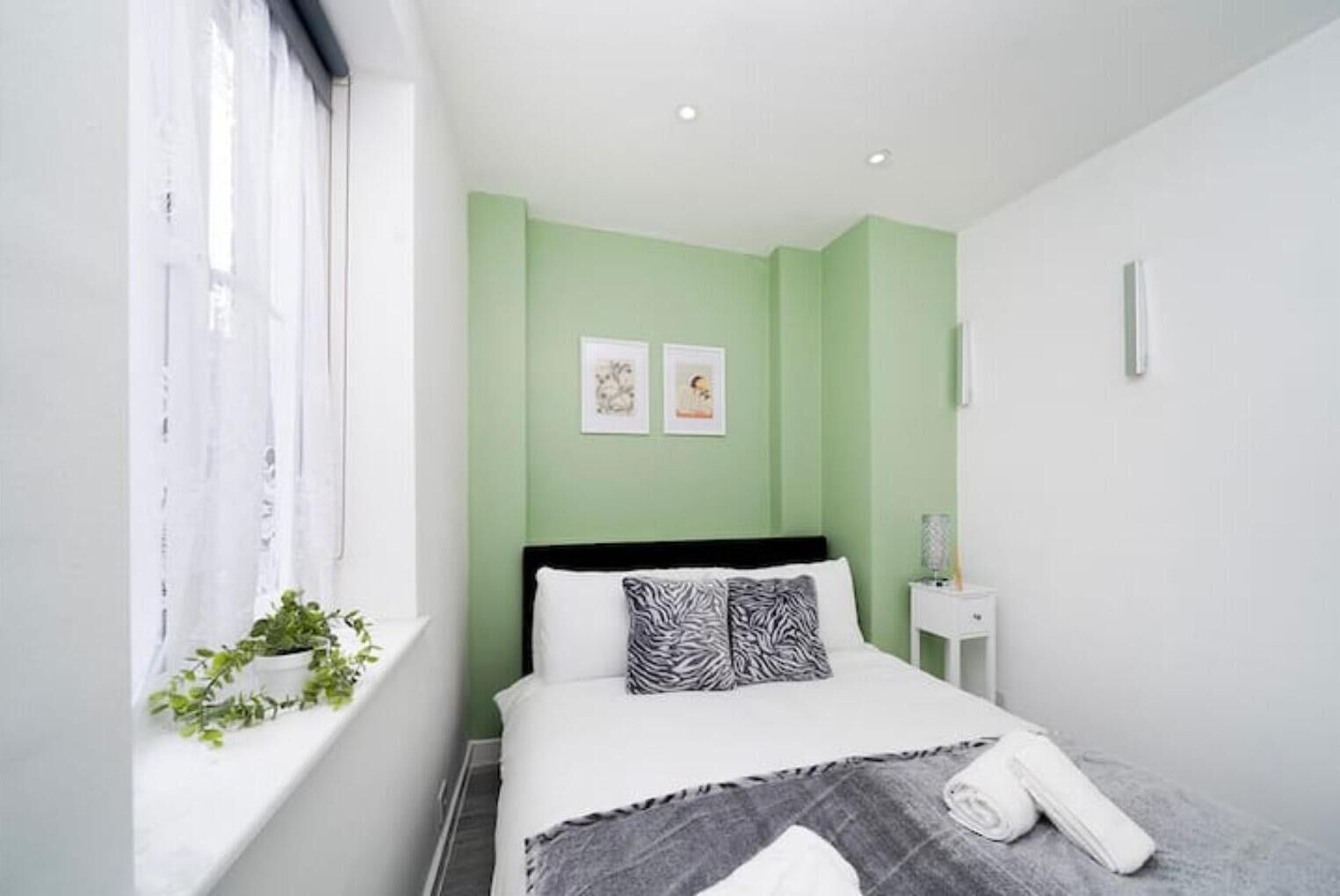 A modern bedroom with a bed, a bedside table, a window with curtains, and a wall-mounted light.