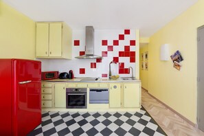 Een compacte keuken met gele kastjes, een rode koelkast, een magnetron, een oven en een spoelbak. De vloer heeft een zwart-wit geruit patroon.