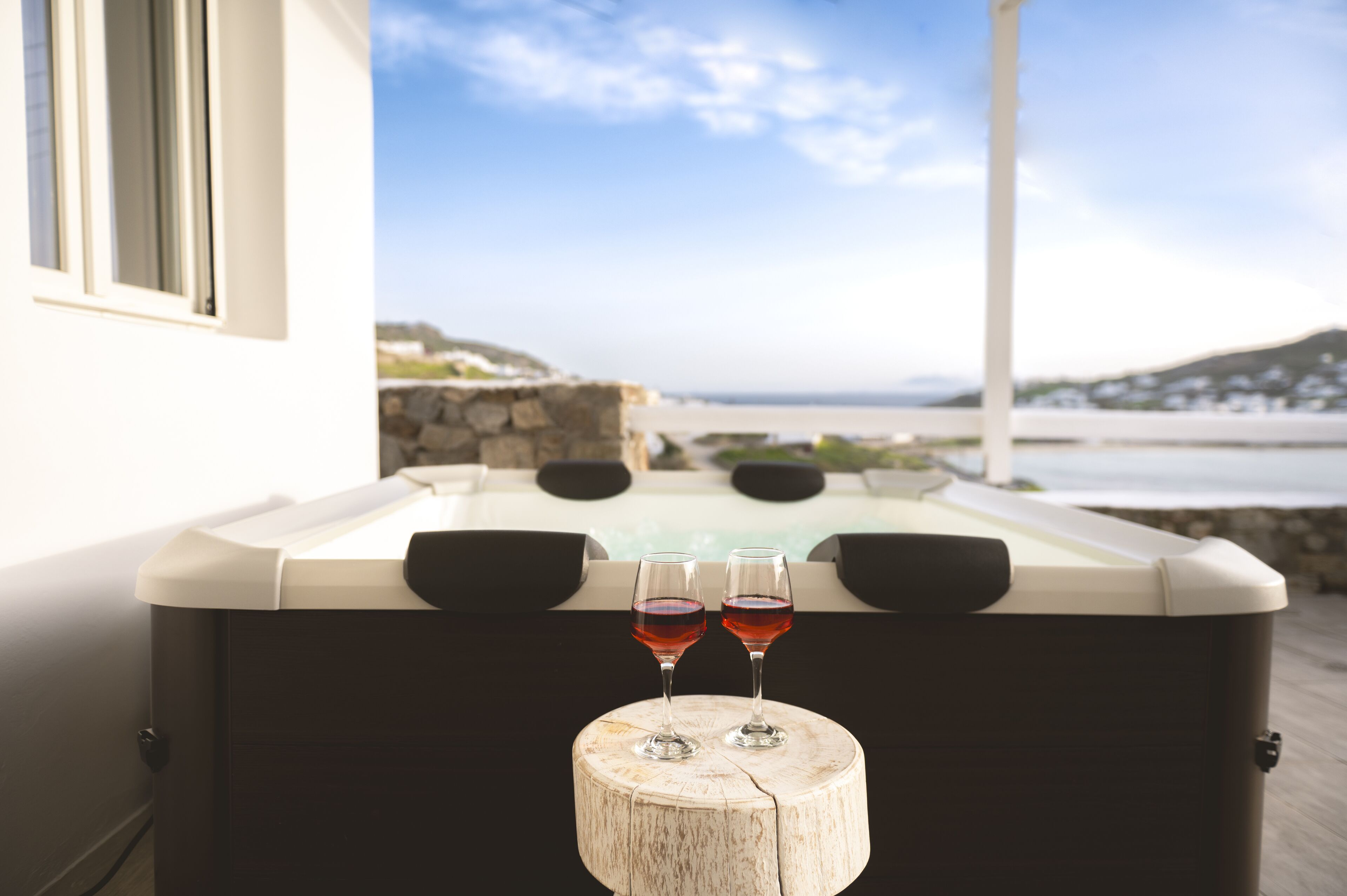 Uma banheira de hidromassagem com duas taças de vinho tinto sobre um banquinho de madeira, com vista para um panorama oceânico.