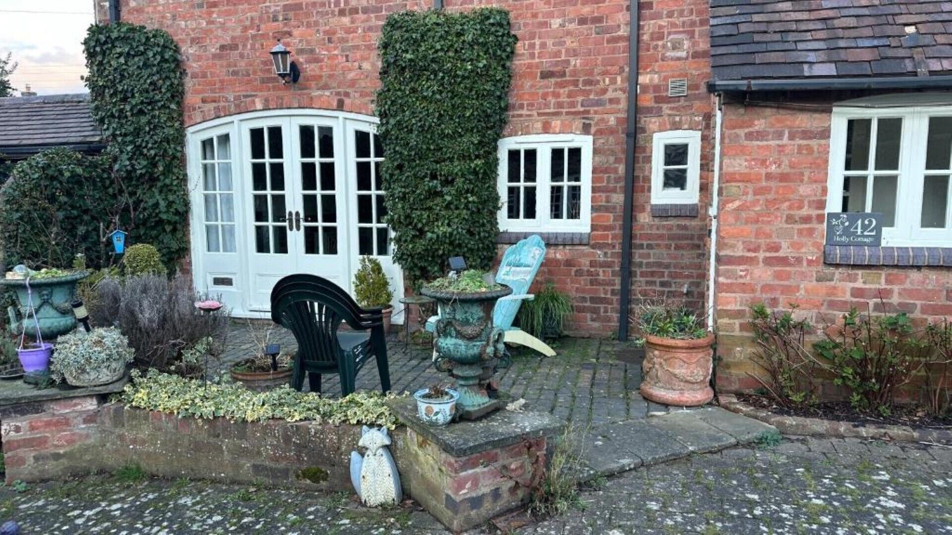 Un bâtiment en briques avec une porte et des fenêtres blanches, entouré de plantes en pot et d’un espace jardin.