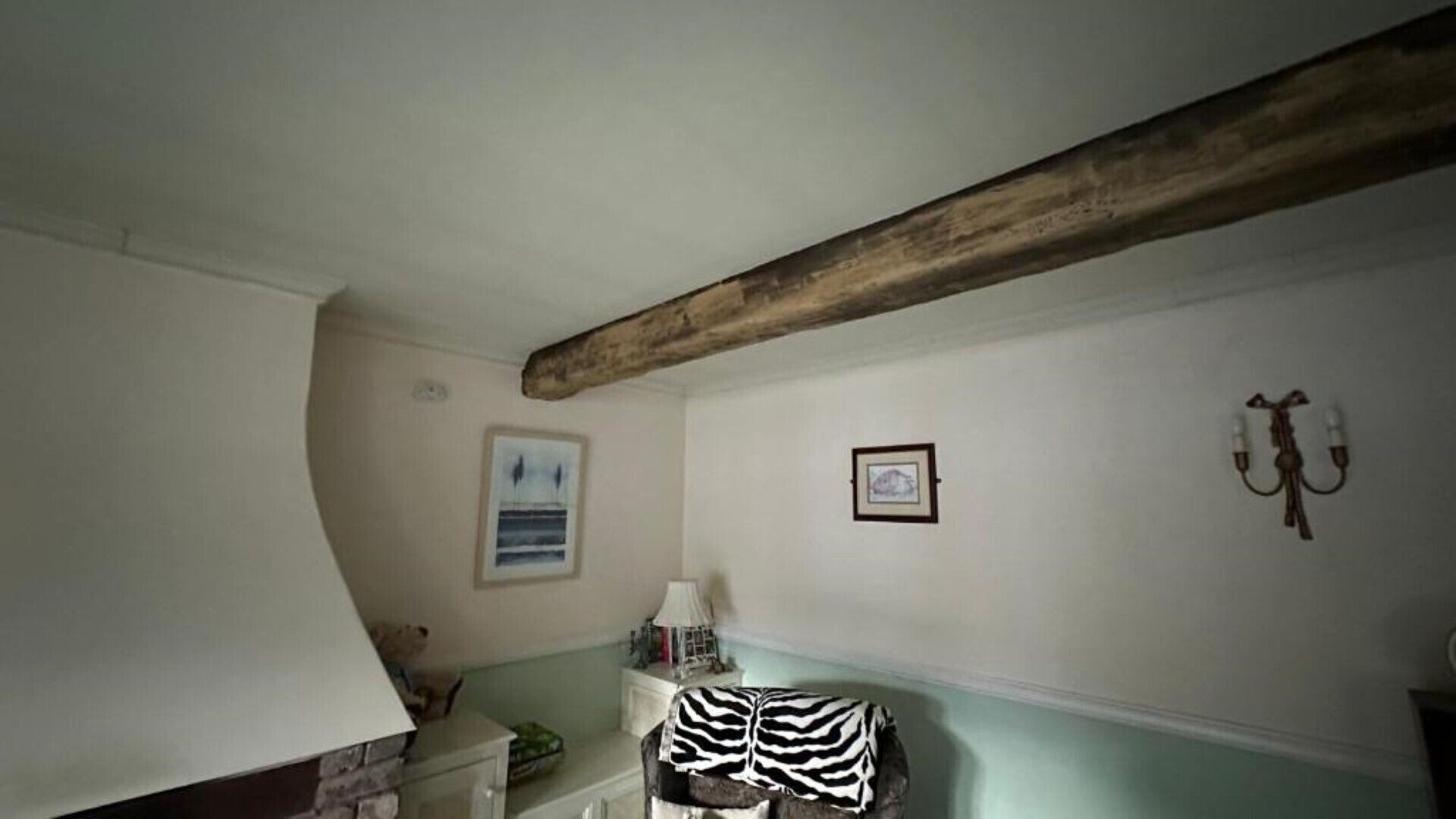 A room with a sloped ceiling, a wooden beam, a zebra-patterned blanket on a chair, a framed picture, and a lamp on a side table.