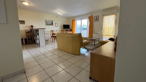 A living room with a sofa, a coffee table, and a TV. There is a kitchen area with a counter and a sink. The room has a tiled floor and a window with curtains.