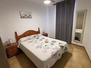 Ein Schlafzimmer mit einem hölzernen Bett, einem Nachttisch mit Lampe, einem Spiegel und einer blumigen Tagesdecke.