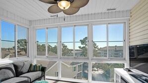 Una habitación luminosa con un gran ventanal, un sofá gris, un otoman turquesa y un mueble blanco para entretenimiento.