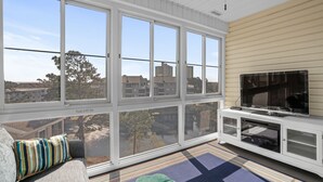 Un balcón con ventanas amplias, una televisión y una zona de estar con cojines.