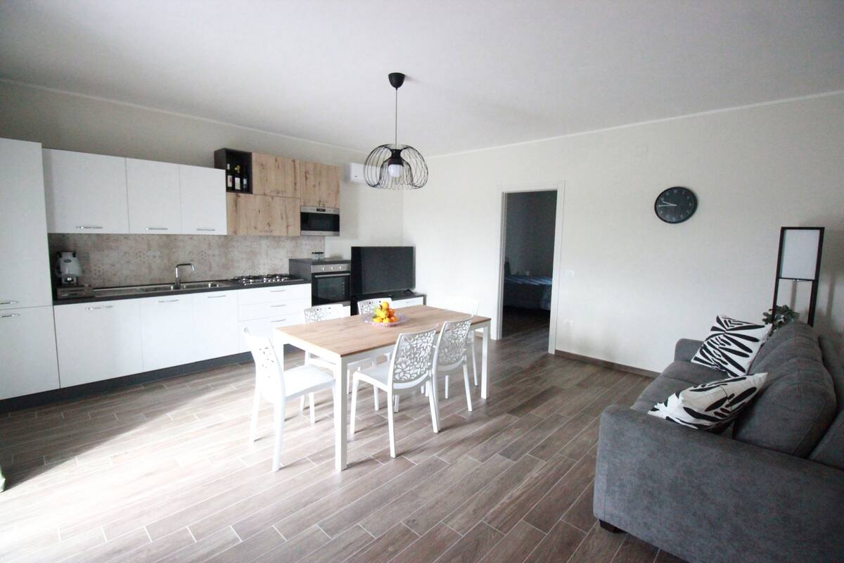 Une cuisine moderne avec des meubles blancs, une table à manger avec des chaises, un canapé gris et une horloge au mur.