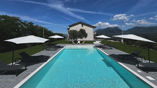 Una piscina con sillas de descanso y sombrillas, rodeada por un edificio y montañas al fondo.
