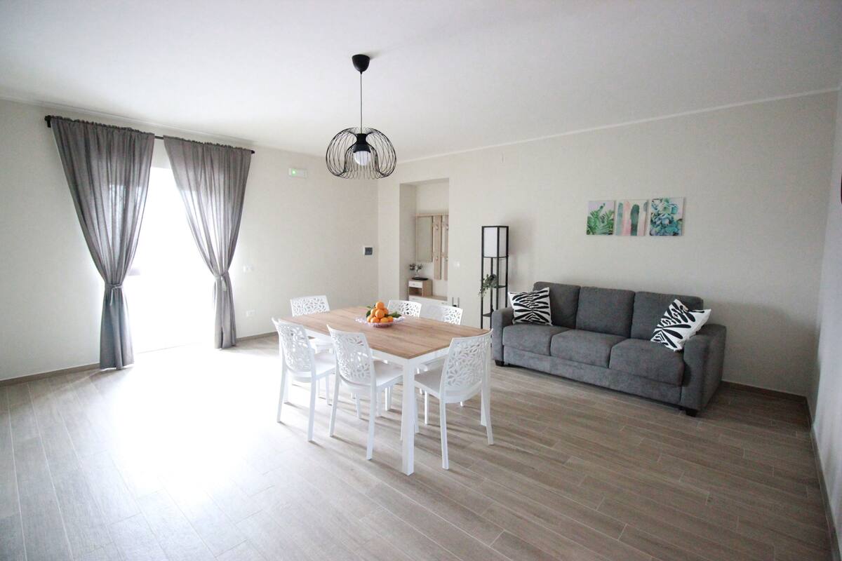 Un salon spacieux comprenant un coin repas, un canapé gris et des chaises blanches.