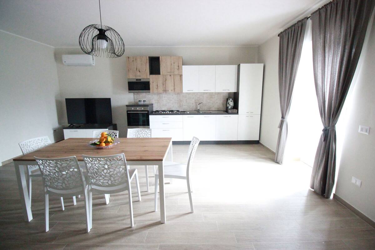 Une cuisine moderne avec des meubles blancs, une table à manger en bois et un téléviseur.