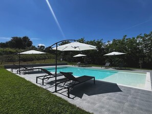 Una piscina con sillones de descanso y sombrillas, rodeada de césped cuidado y árboles.