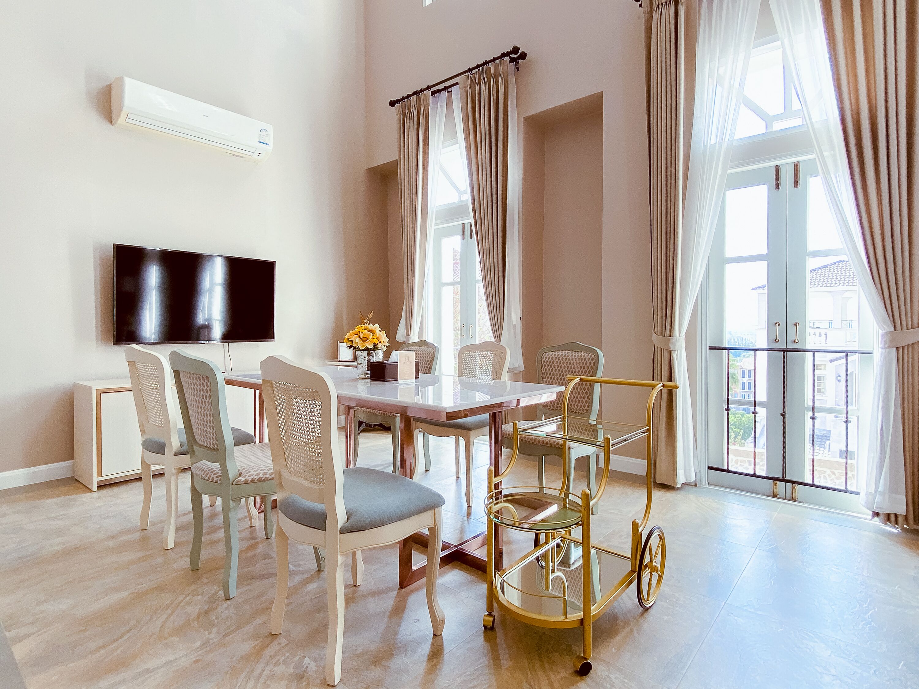 A modern dining area with a glass table, cushioned chairs, a gold-framed cart, and a flat-screen TV mounted on the wall.