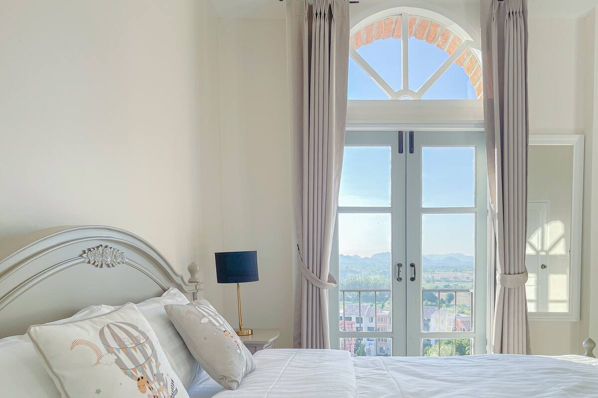 A bedroom with a large bed, a nightstand, a lamp, and a view of the city through a large window.