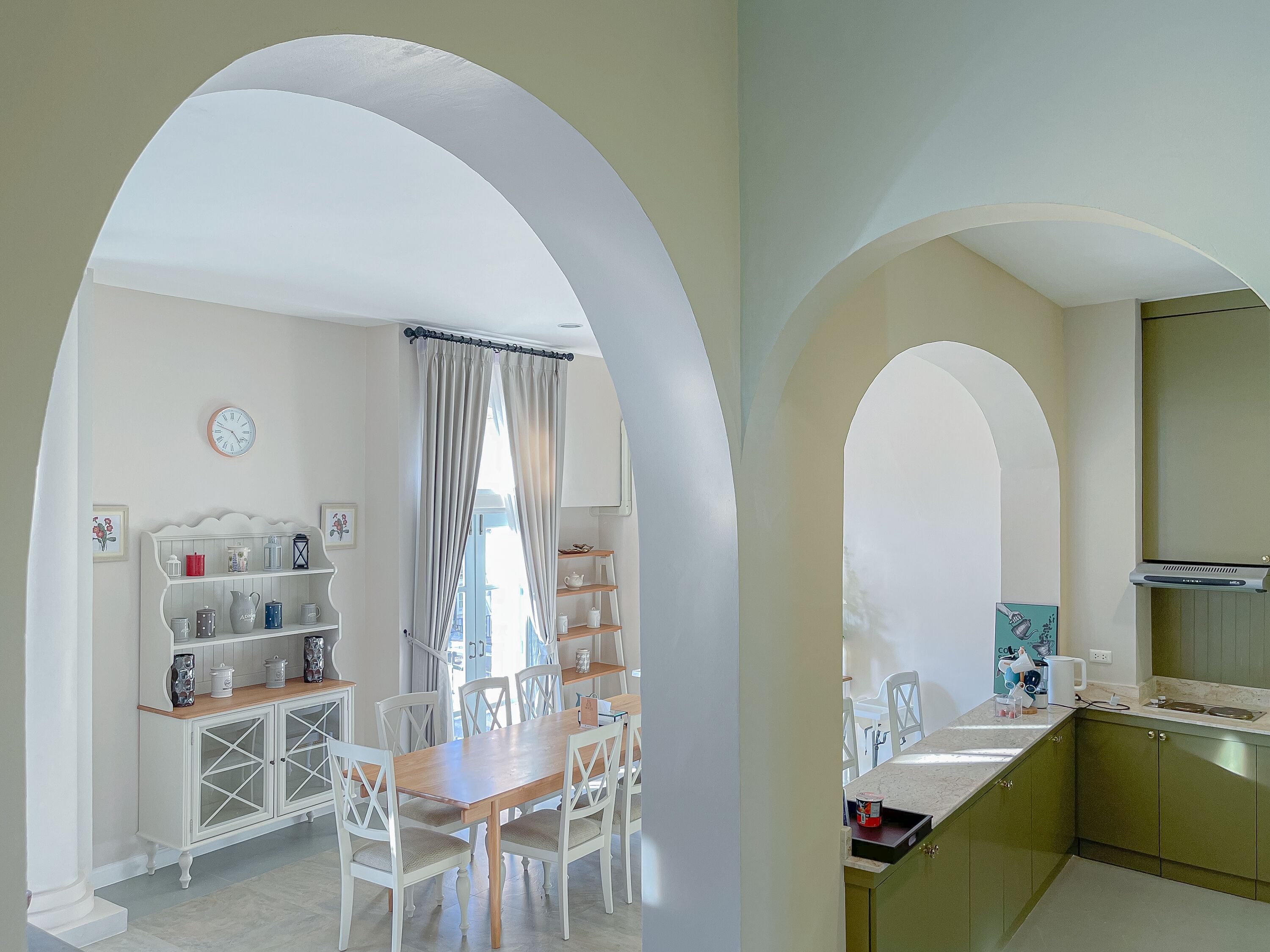 Un comedor con una mesa y sillas de madera, un aparador blanco con estantes, y una cocina con armarios verdes y una encimera.