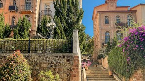 Un edificio histórico con ventanales de arco, una torre, abundante vegetación y una escalera de piedra.