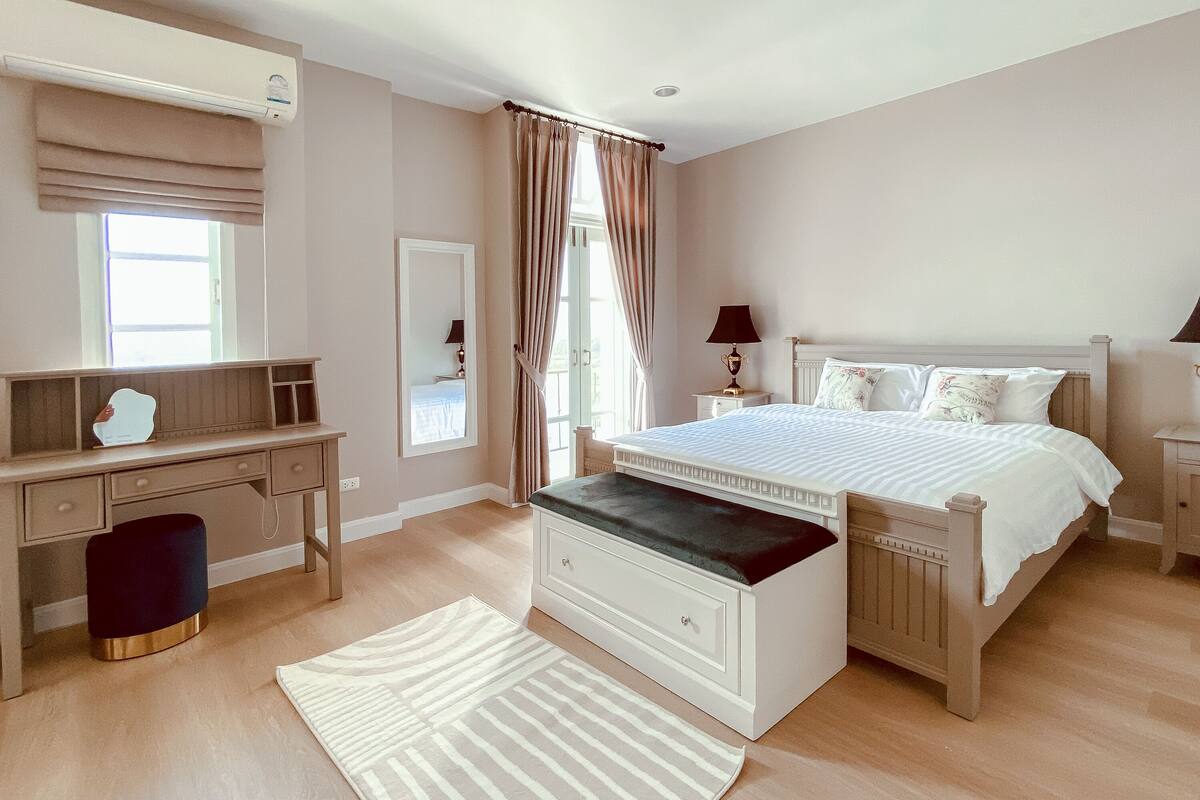 A bedroom with a bed, a desk, a chair, a bench, a mirror, and a window.