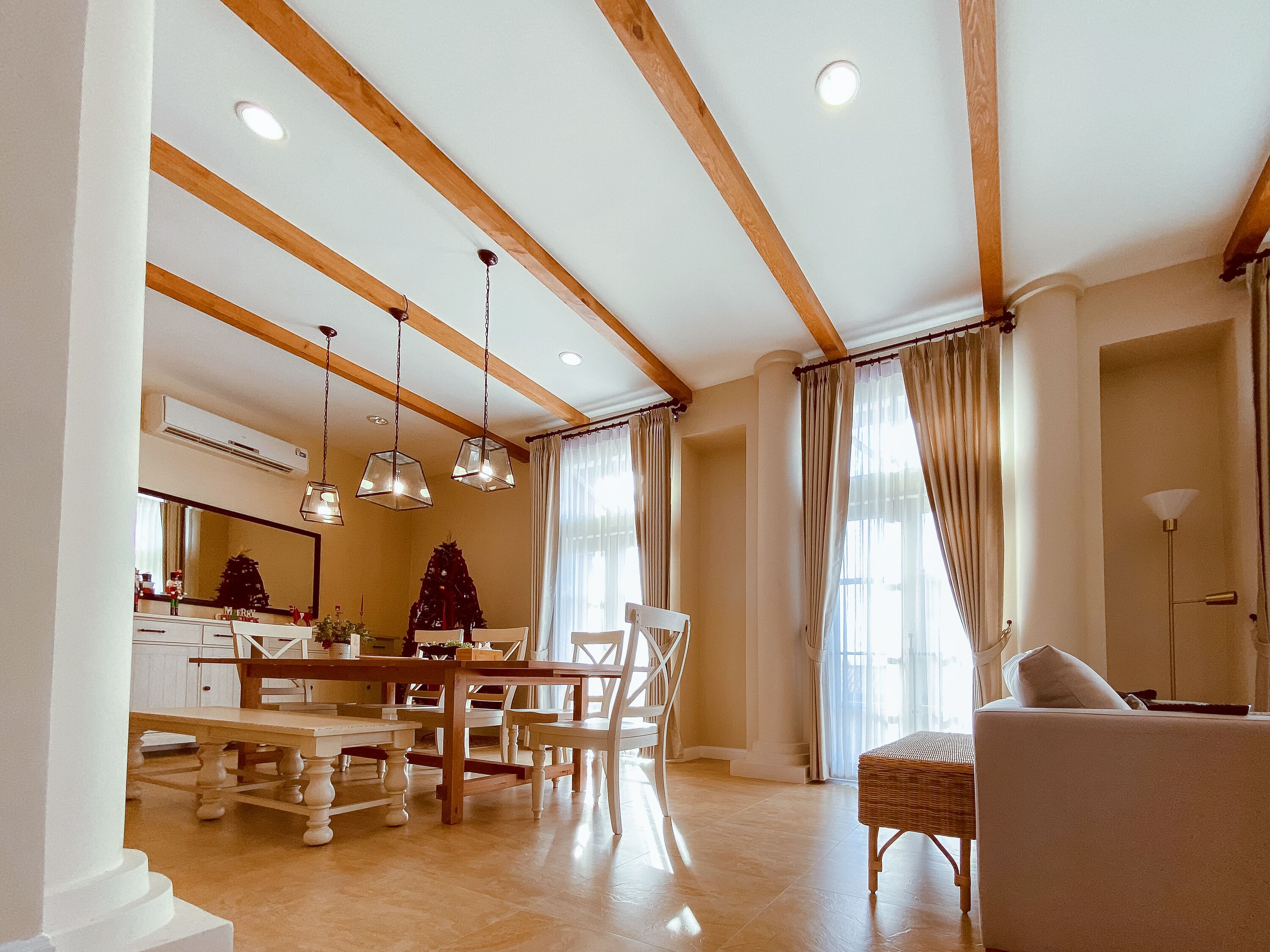 A spacious living area with a wooden ceiling, a dining table, chairs, a sofa, and a large window with curtains.