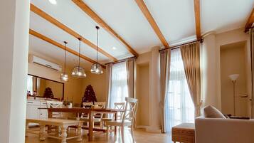 A spacious living area with a wooden ceiling, a dining table, chairs, a sofa, and a large window with curtains.