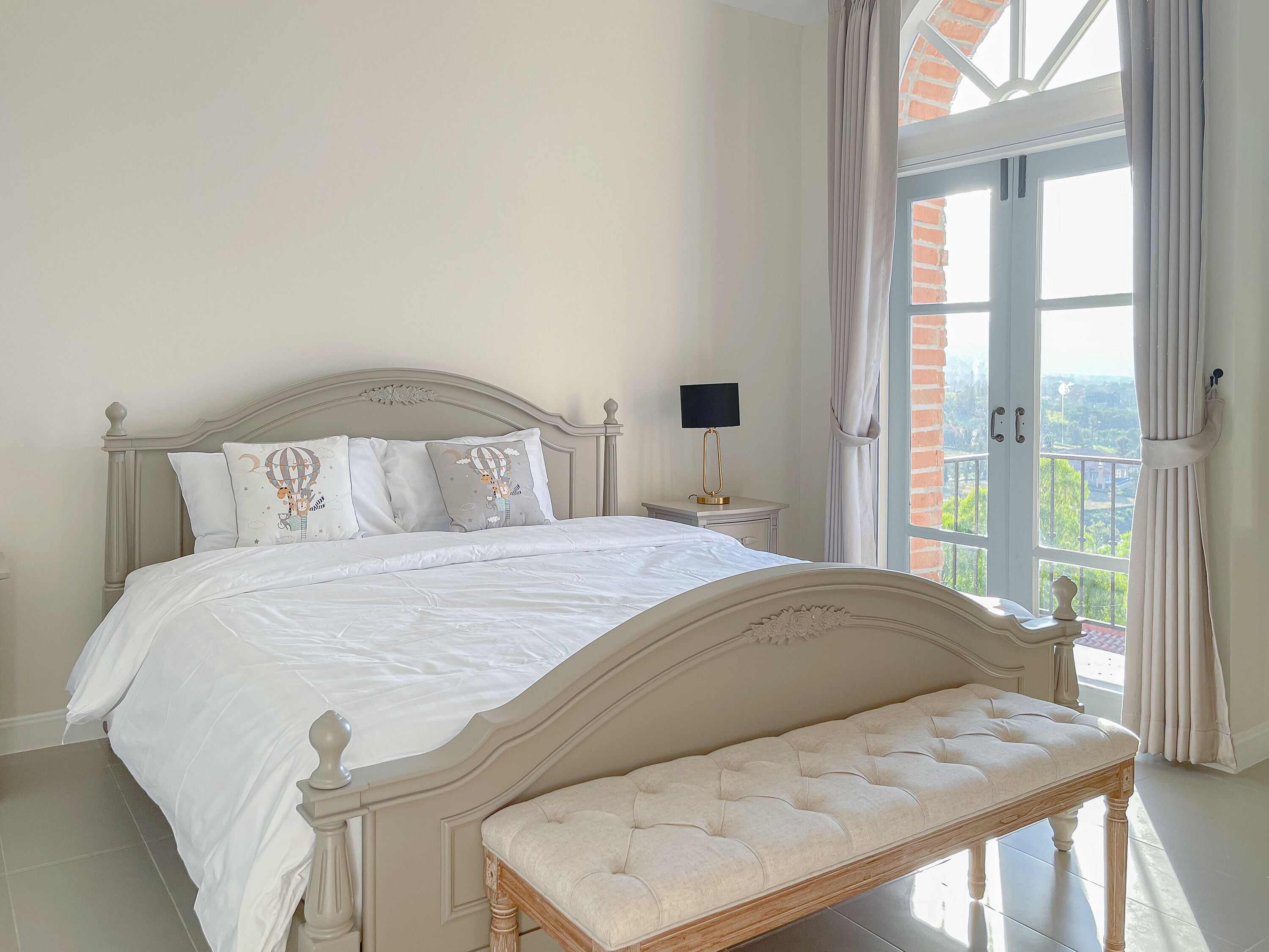 A bedroom with a large bed, a bench, a nightstand, and a view of greenery through the window.
