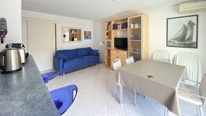 Un espacio habitable compacto con kitchenette, un sofá azul, una mesa de comedor con sillas y una estantería.