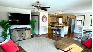 A spacious living room with a large flat-screen TV, a ceiling fan, and a kitchen in the background. - Orvis Outpost
 (Ridgway)