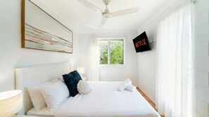 Ein Hotelzimmer mit Bett, Deckenventilator, an der Wand befestigtem Fernseher und einem Fenster mit Blick auf Grünflächen.
