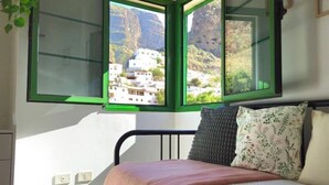 A bedroom with a bed, a window with a view of buildings and mountains, and a green-framed window. - La Villa del Valle (CN)