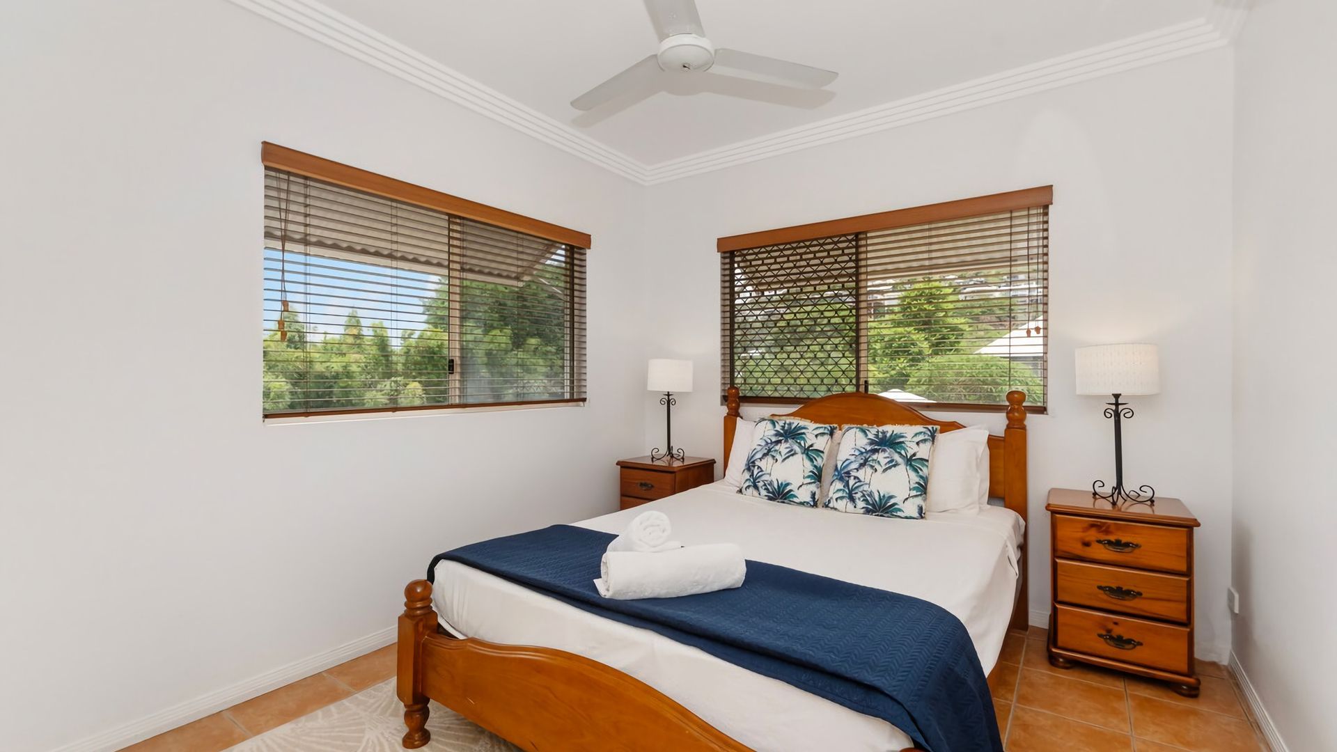 A bedroom with a bed, two nightstands, a ceiling fan, and windows with blinds.