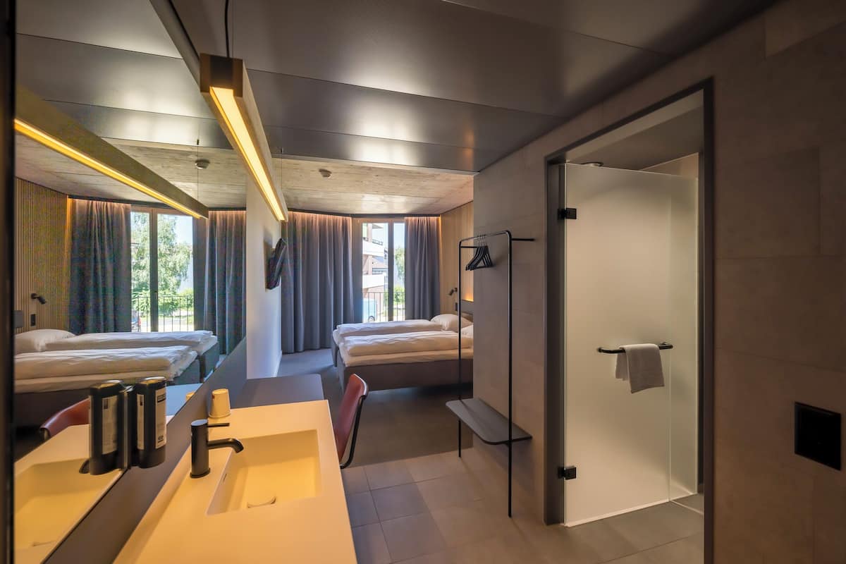 Una habitación de hotel moderna con una cama grande, zona de ducha separada, tocador con espejo y vistas al exterior a través de amplias ventanas.