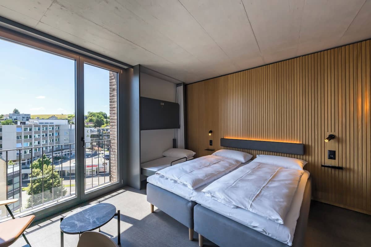 Una habitación de hotel moderna con un ventanal grande, una cama, una mesita pequeña y un rincón de descanso.